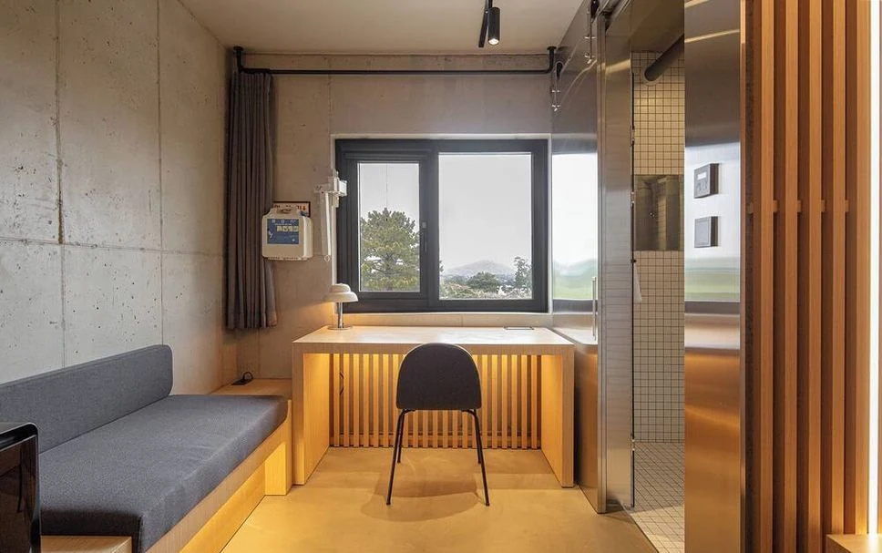 Compact guest room at Playce Camp Jeju showing desk, window and bathroom layout