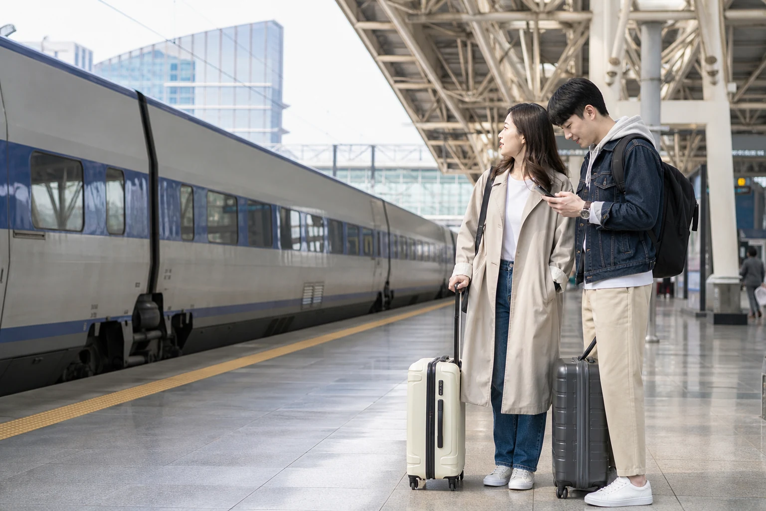 A traveler checks their phone on a cross-city train platform in South Korea with luggage in hand
