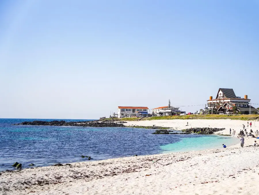 Jeju Udo Rhodolith Beach with pale shore particles, blue-green water and coastal buildings